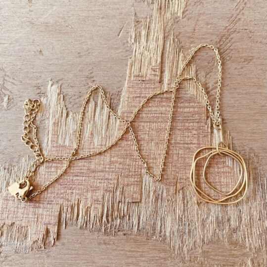 Collier chaîne fine dorée avec pendentif de trois anneaux irréguliers entrelacés, posé sur bois,