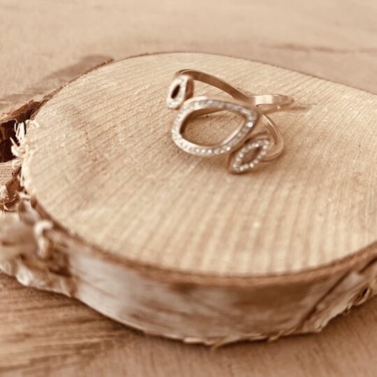 Bague dorée rosée avec trois formes ajourées serties de petits strass, posée sur rondin de bois, Calina