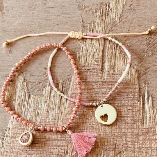 Deux bracelets corail et dorés portés au poignet, avec pampille rose, breloque scintillante et médaille cœur ajouré